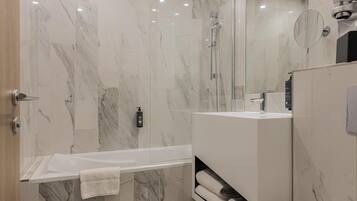 Een moderne badkamer met een marmeren vloer, een douche met glazen wand, een wit bad en een ingebouwde wastafel met spiegel.