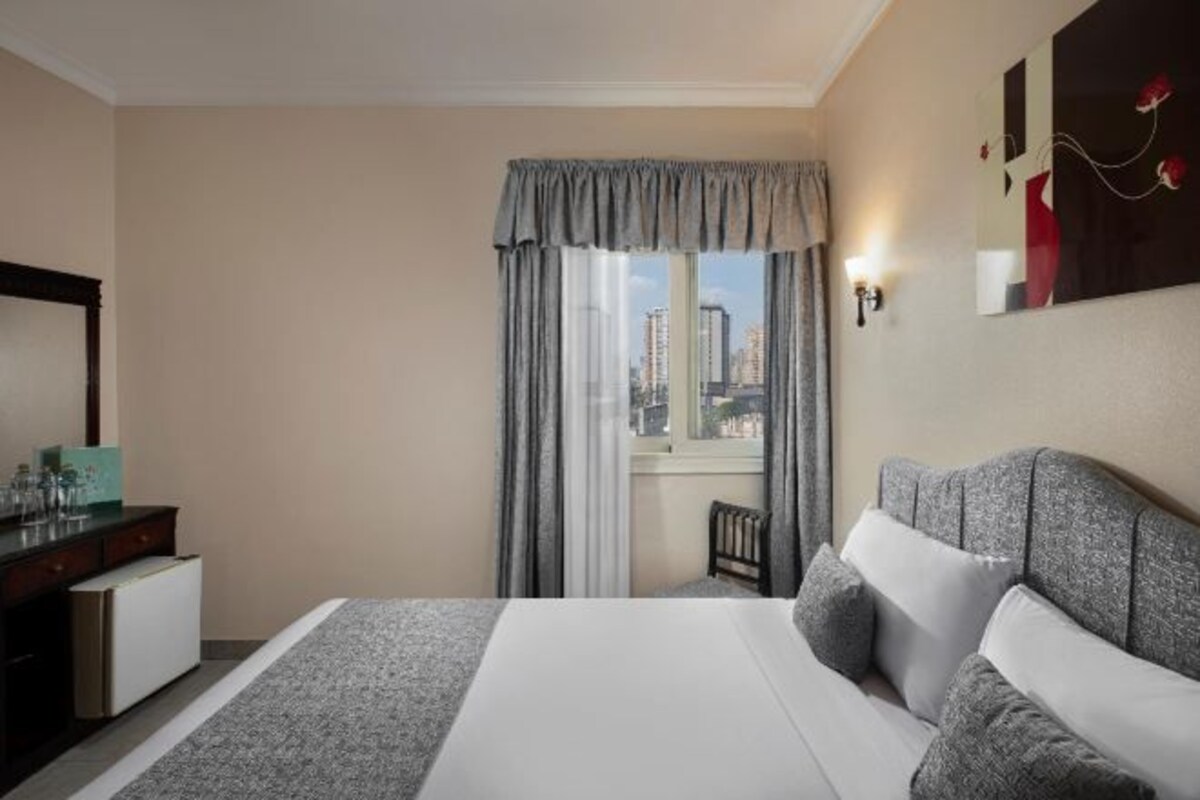 Habitación de hotel con cama, escritorio, silla, ventana con cortinas y vista a la ciudad.