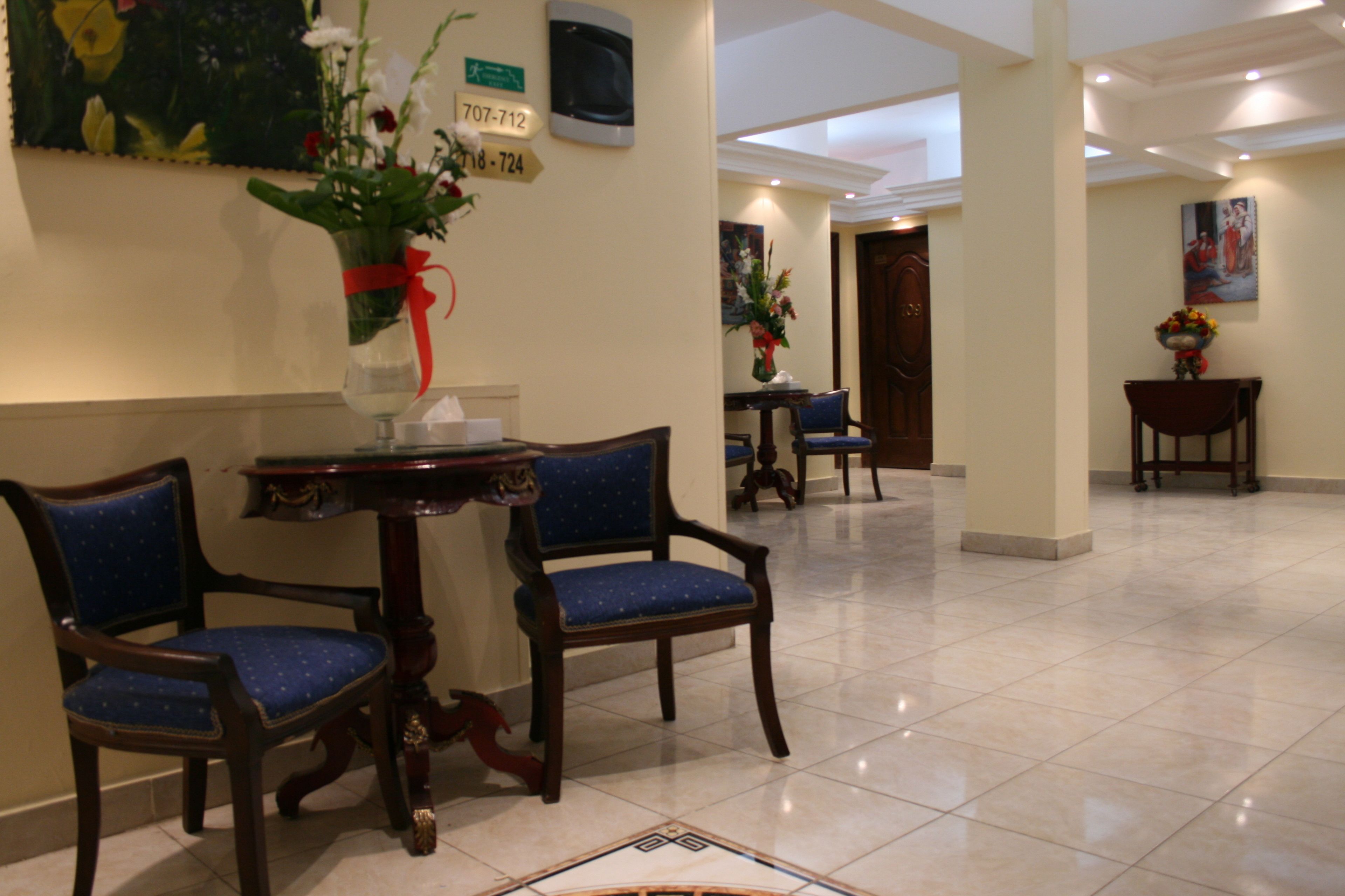 A hotel lobby with a reception desk, two blue upholstered chairs, a wooden table, and a tiled floor.