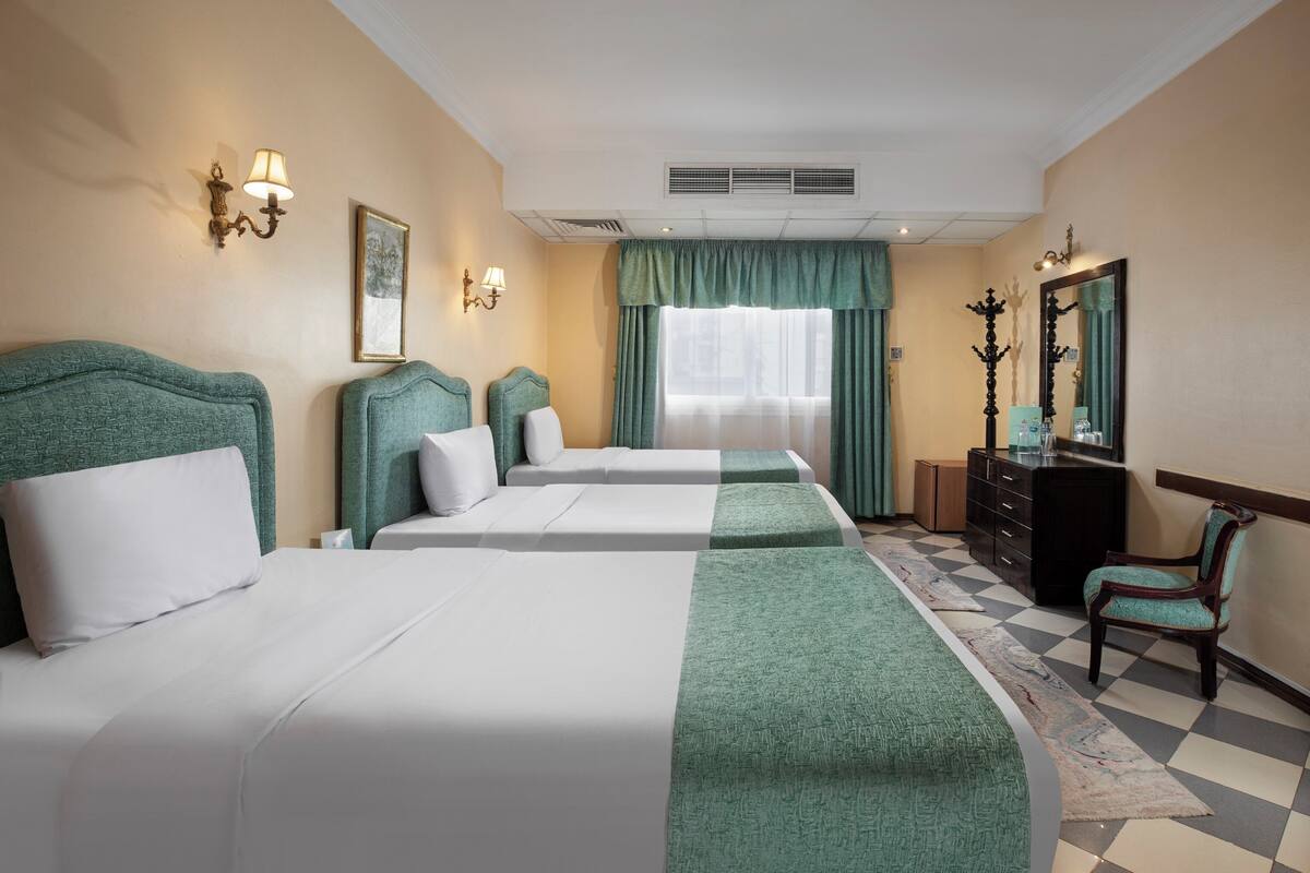 Quarto de hotel com duas camas, uma cômoda, uma cadeira, um espelho e uma janela com cortinas.