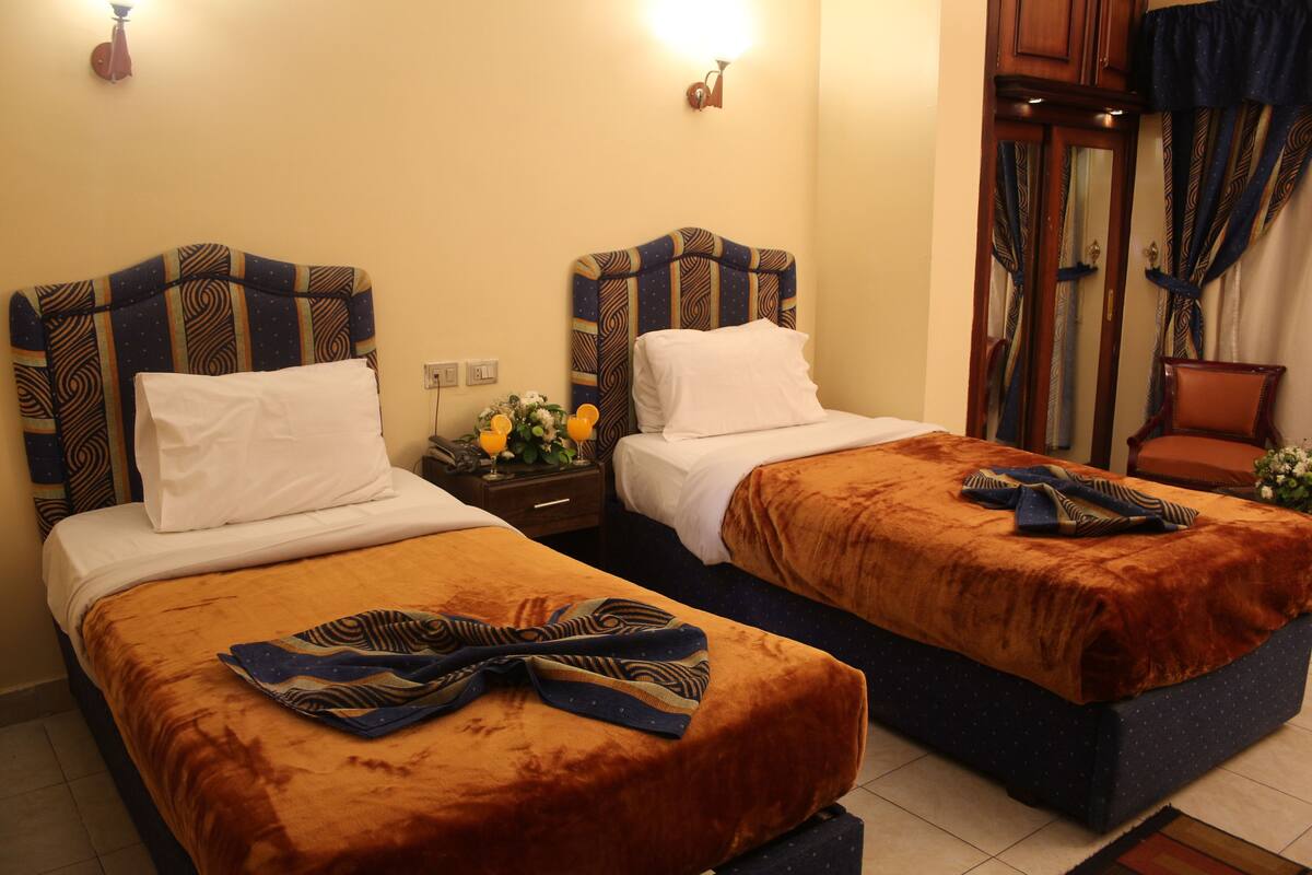 Quarto de hotel com duas camas, cada uma com cabeceira estampada, almofadas brancas e manta marrom. Tem uma mesa de cabeceira com abajur e um vaso de flores.