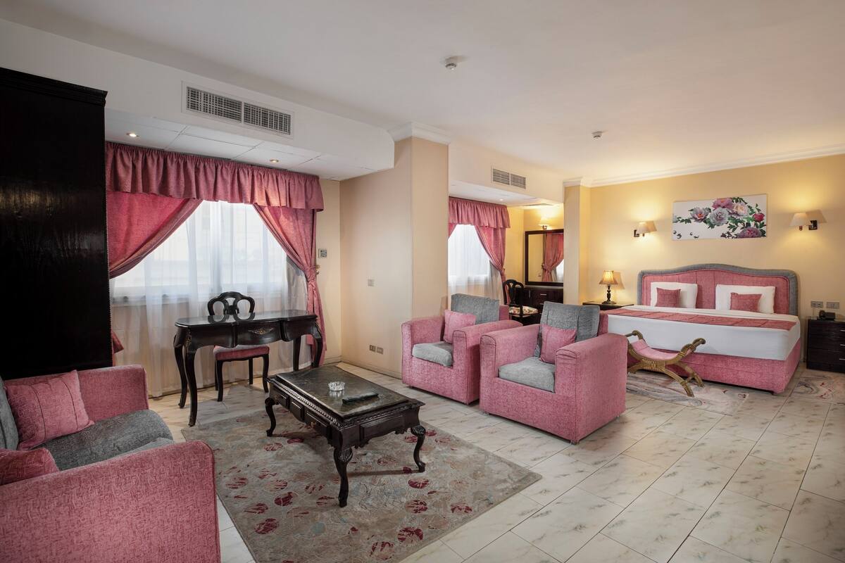 Quarto de hotel com cama, duas poltronas cor-de-rosa, mesa de centro, mesa de jantar com cadeiras e um espelho grande.