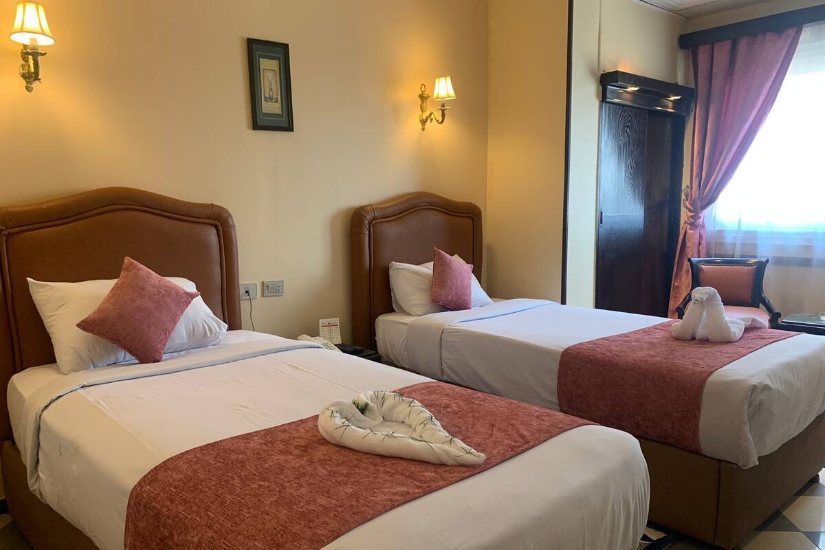 Duas camas de casal com cabeceiras castanhas, roupa de cama branca e almofadas cor-de-rosa. Há arandelas com luminárias, um quadro emoldurado e uma cadeira com uma toalha branca.