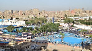 Une grande piscine extérieure entourée de chaises longues bleues et de parasols, avec une vue sur la ville en arrière-plan.