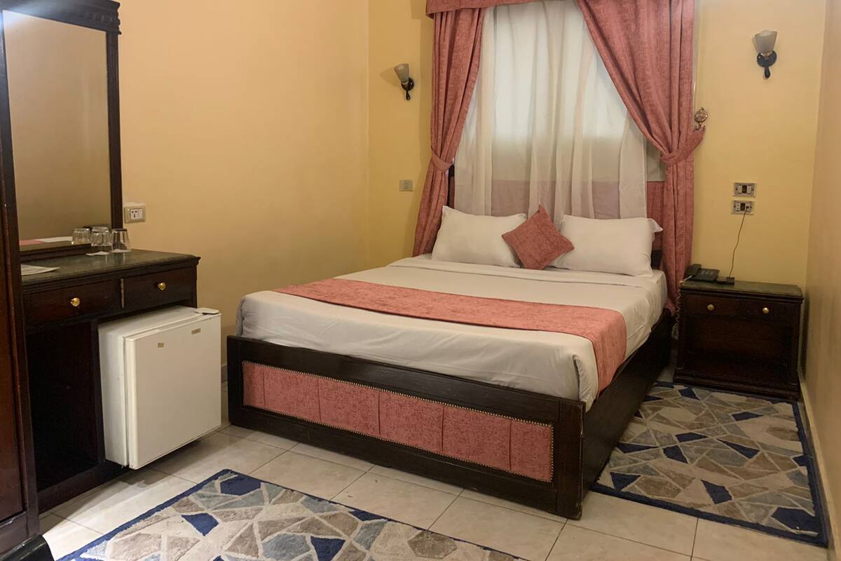 Quarto com uma cama, um frigorífico pequeno branco, uma cómoda de madeira, uma mesa de cabeceira e um espelho.
