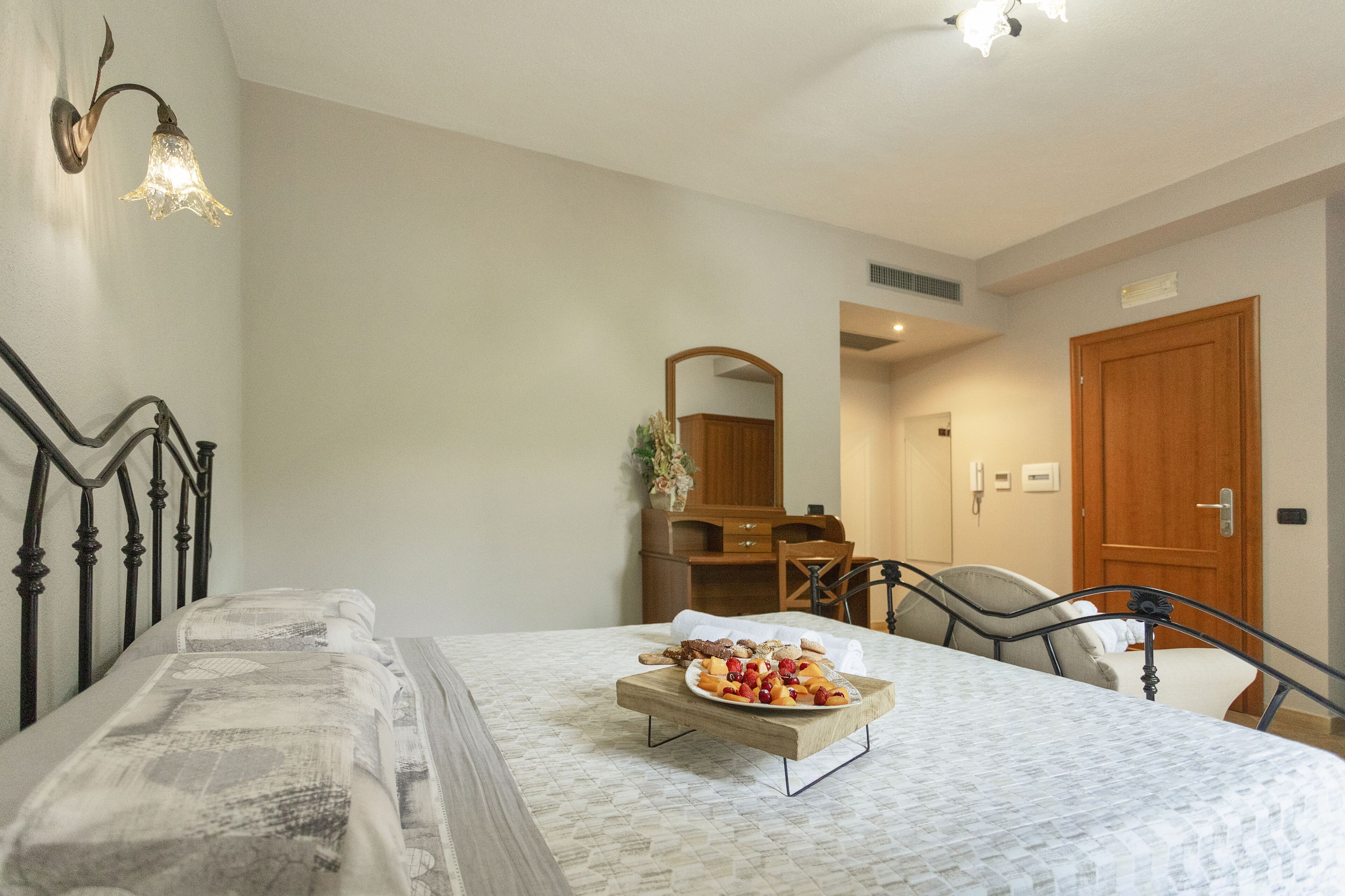 Un dormitorio con cama, mesita de noche con una bandeja de frutas, cómoda, espejo y puerta.