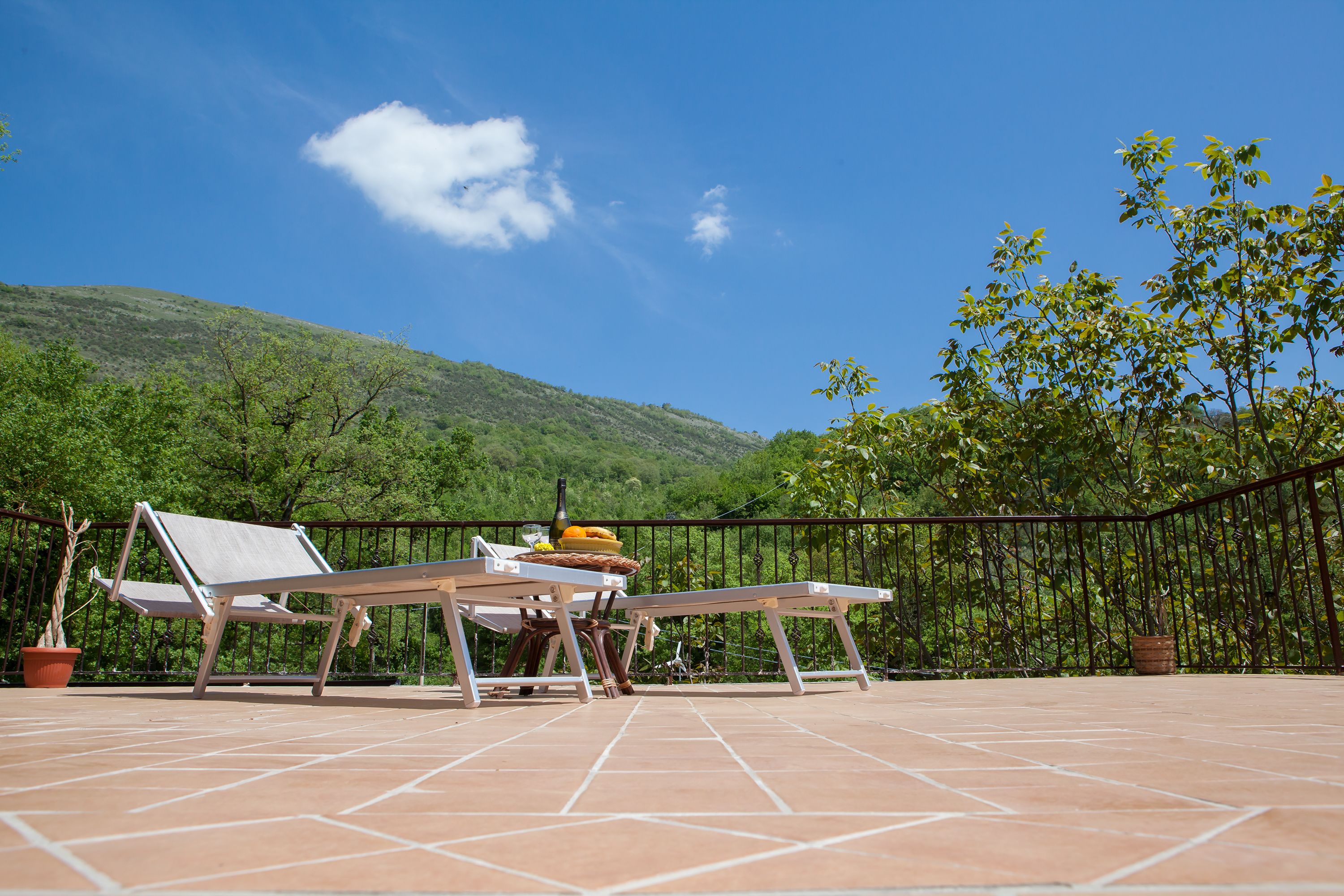 An outdoor terrace with two lounge chairs, a table with a bottle and fruit, and a view of a green hillside.