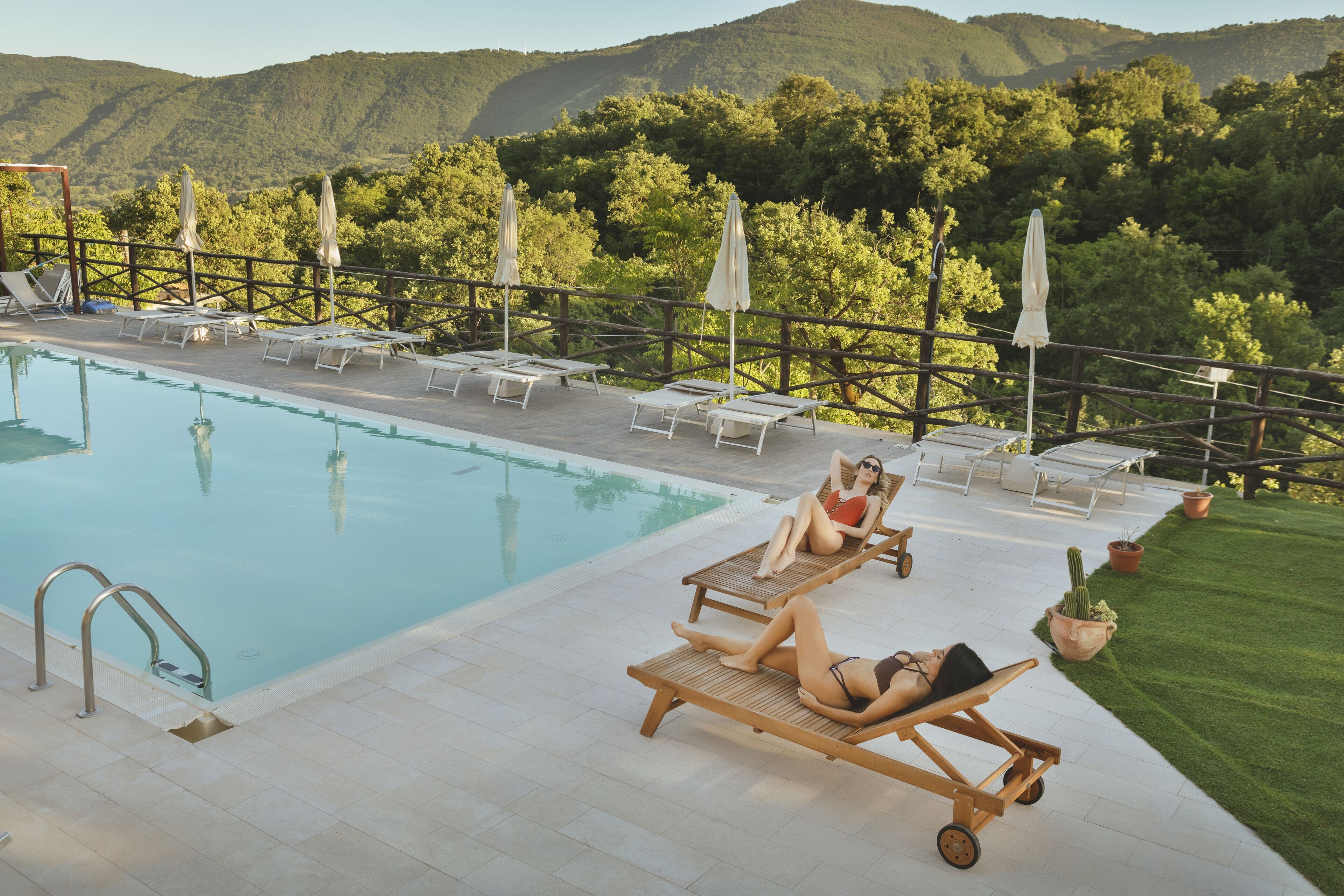 A pool area with lounge chairs and umbrellas, surrounded by a wooden fence and lush greenery.