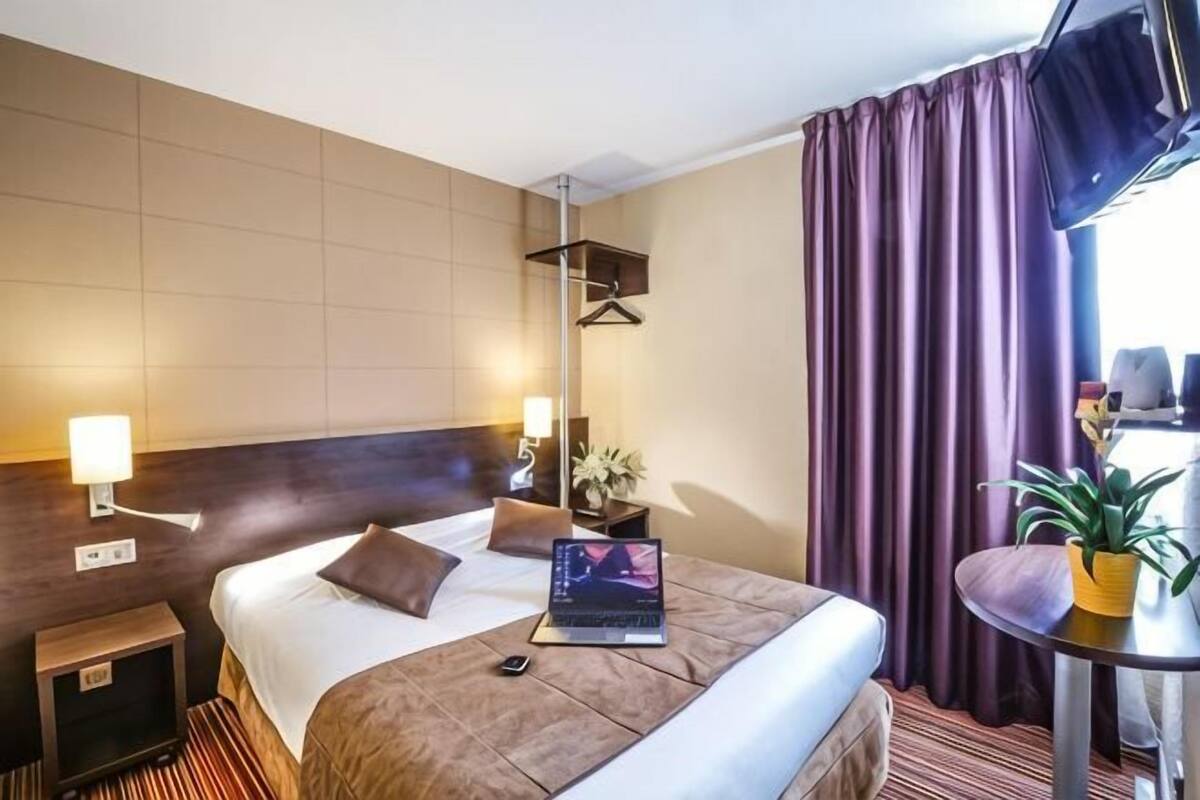 A hotel room with a bed, bedside lamps, a laptop, a small table with a plant, and a window with curtains.