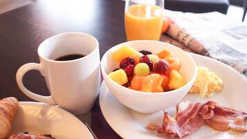 A breakfast setting with a cup of coffee, a bowl of fruit, scrambled eggs, bacon, and croissants on a table.