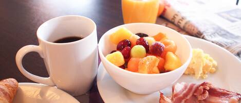 A breakfast setting with a cup of coffee, a bowl of fruit, scrambled eggs, bacon, and croissants on a table.