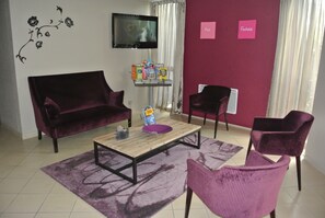 A living room with a dark purple sofa, a wooden coffee table, and two matching armchairs. A flat-screen TV is mounted on the wall, and there are two pink signs on the wall. - Hôtel Inn Design & Restaurant Le Mans (Arnage)