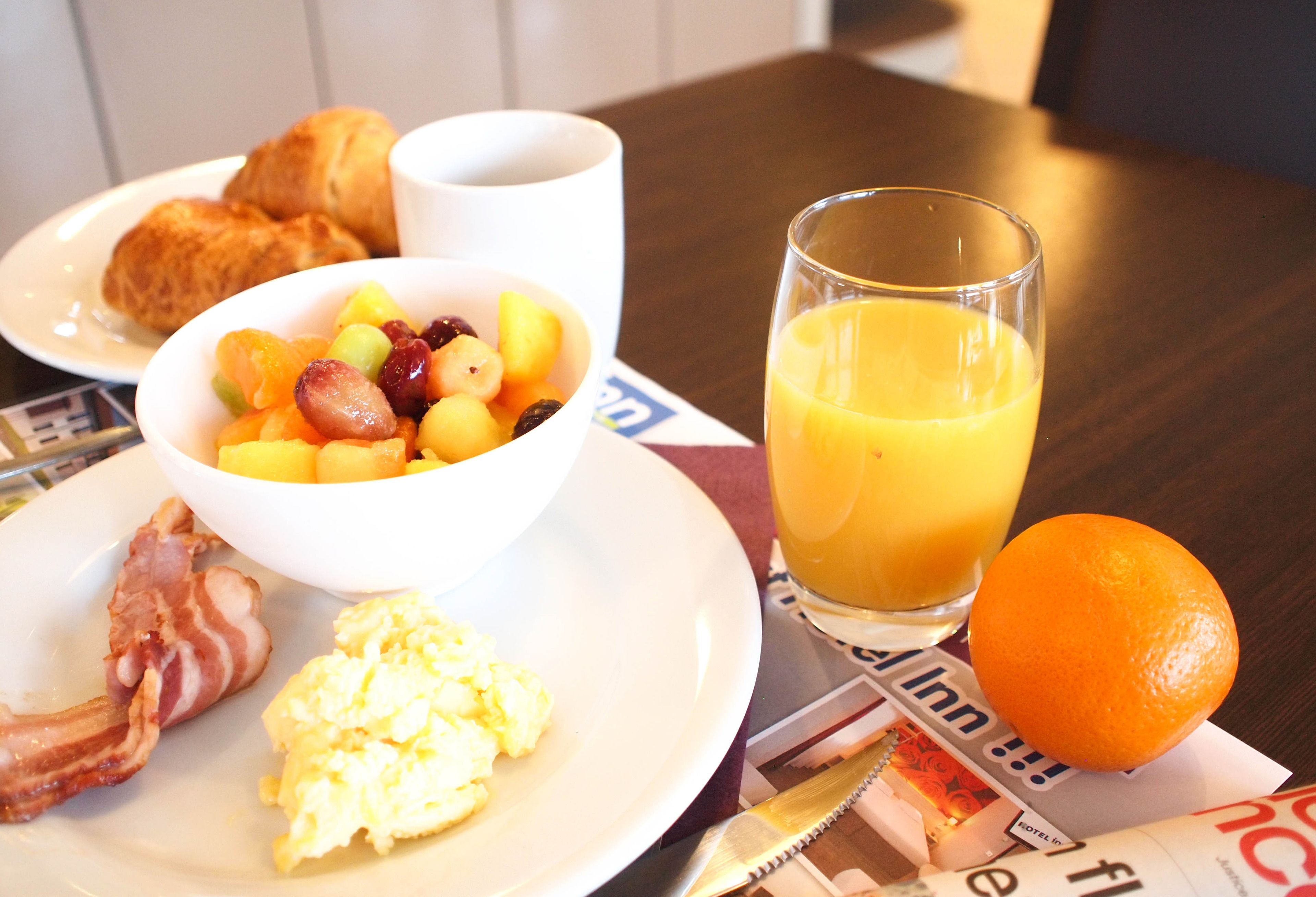 Ein Teller mit Frühstücksei, Speck, Obst und einem Glas Orangensaft auf einem Tisch mit Zeitungen.