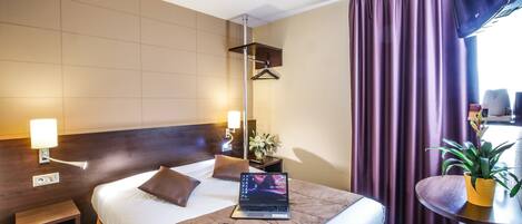 A hotel room with a bed, bedside table, lamp, laptop, and a small table with a plant.