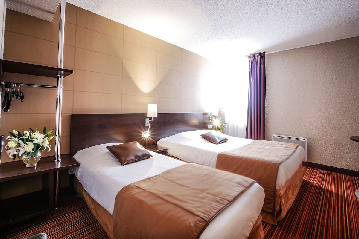 A hotel room with two beds, a desk with a lamp, and a vase of flowers.