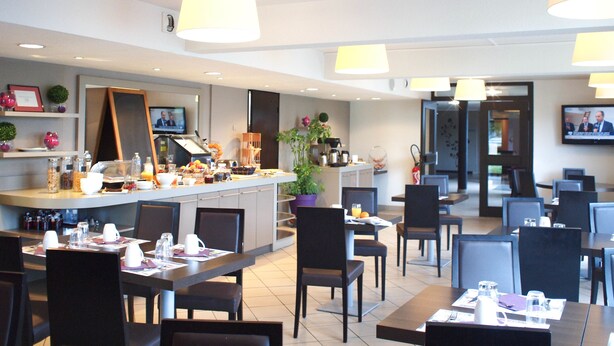 A hotel dining area with multiple tables and chairs, a kitchen counter with various items, and a television screen displaying a news program. - Hôtel Inn Design & Restaurant Le Mans (Arnage)