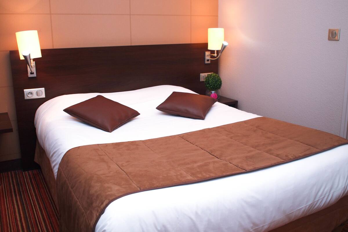 A neatly made bed with a brown headboard, two bedside lamps, and a small potted plant on the nightstand.