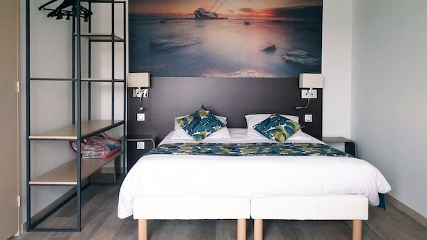 Ein Schlafzimmer mit einem Bett, zwei Kissen, einem Nachttisch, einer Wandlampe, einem Hakenleistenregal und einem großen Gemälde, das einen Sonnenuntergang über Wasser zeigt.