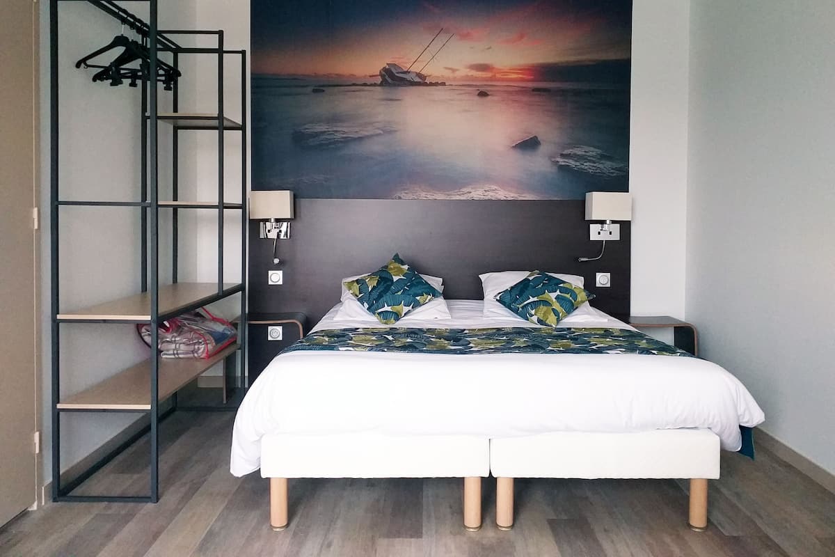 Une chambre à coucher comprenant un lit, deux oreillers, une table de chevet, une lampe fixée au mur, une étagère avec des cintres et un grand tableau représentant un coucher de soleil sur l’eau.