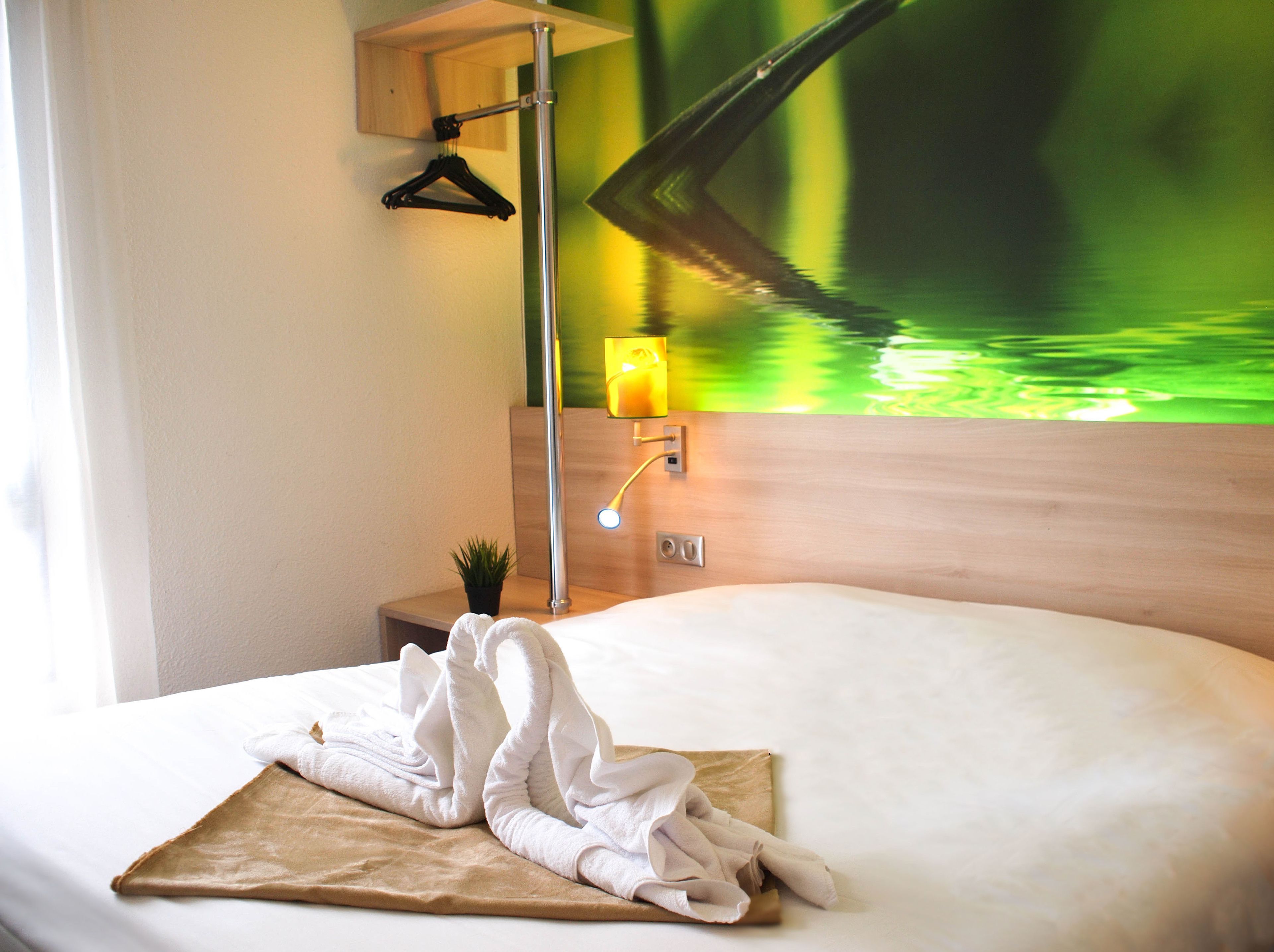 A hotel room with a bed, a towel, a small plant, and a wall-mounted lamp.