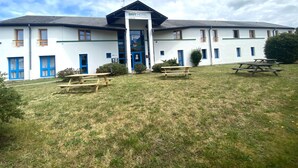Ein Hotel mit weißer Fassade, blauen Türen und einem überdachten Eingangsbereich. Auf der Rasenfläche vor dem Haus stehen Picknicktische.