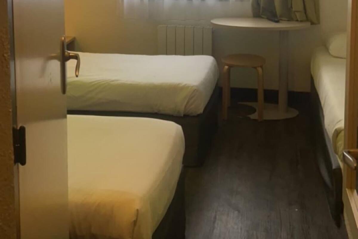 Une petite pièce, de taille modeste, comprenant deux lits simples, un petit bureau et une fenêtre ornée de rideaux légers.