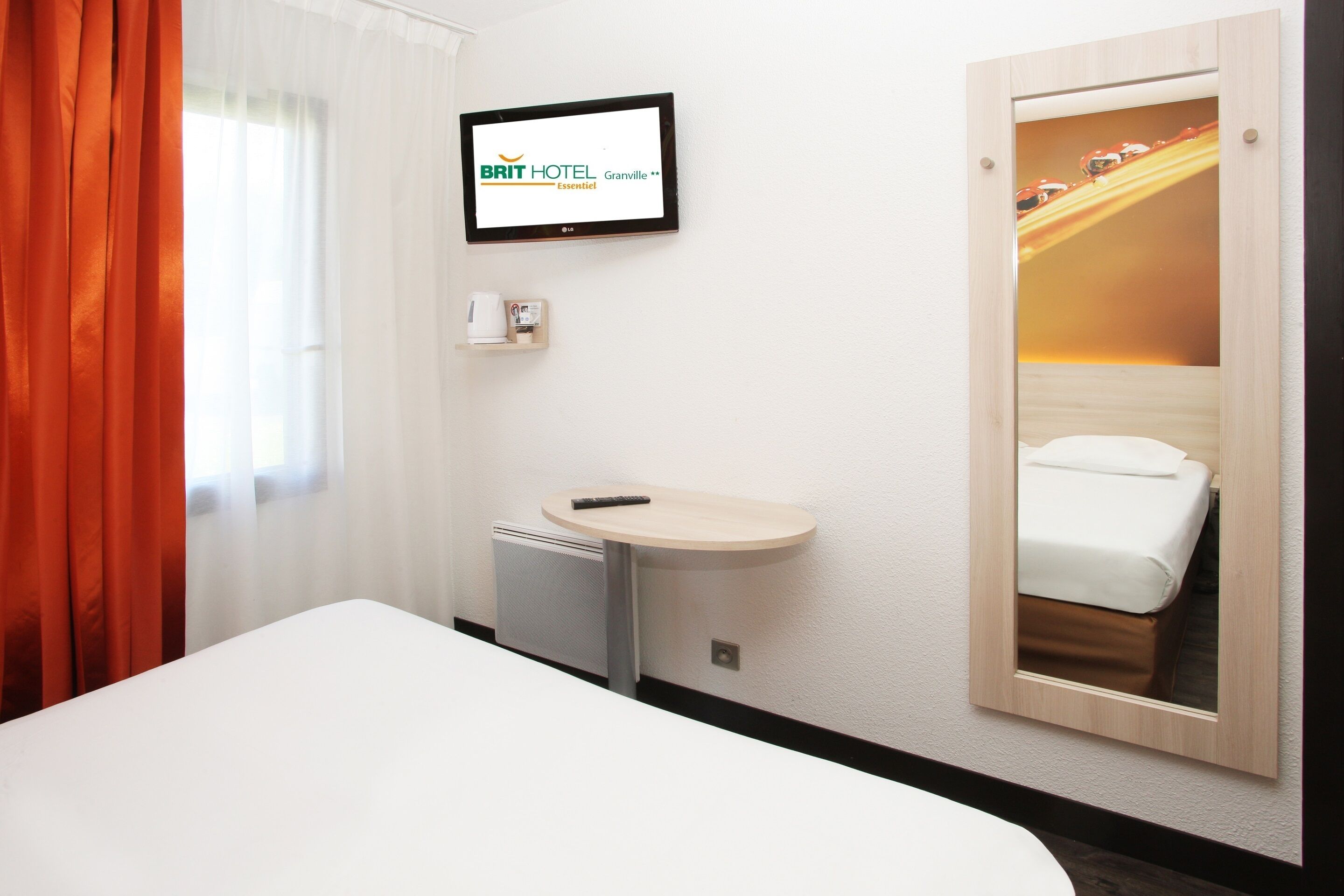 A hotel room with a bed, a small desk with a remote control, a television mounted on the wall, and a mirror reflecting the bed.