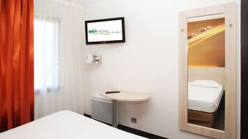 A hotel room with a bed, a small desk with a remote control, a television mounted on the wall, and a mirror reflecting the bed.