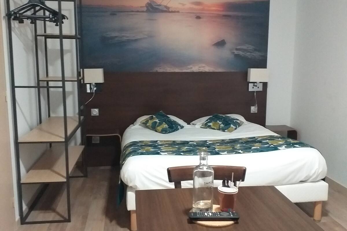 Une chambre d’hôtel comprenant un lit, un bureau, une chaise, une étagère et un grand tableau mural représentant un coucher de soleil sur l’eau.