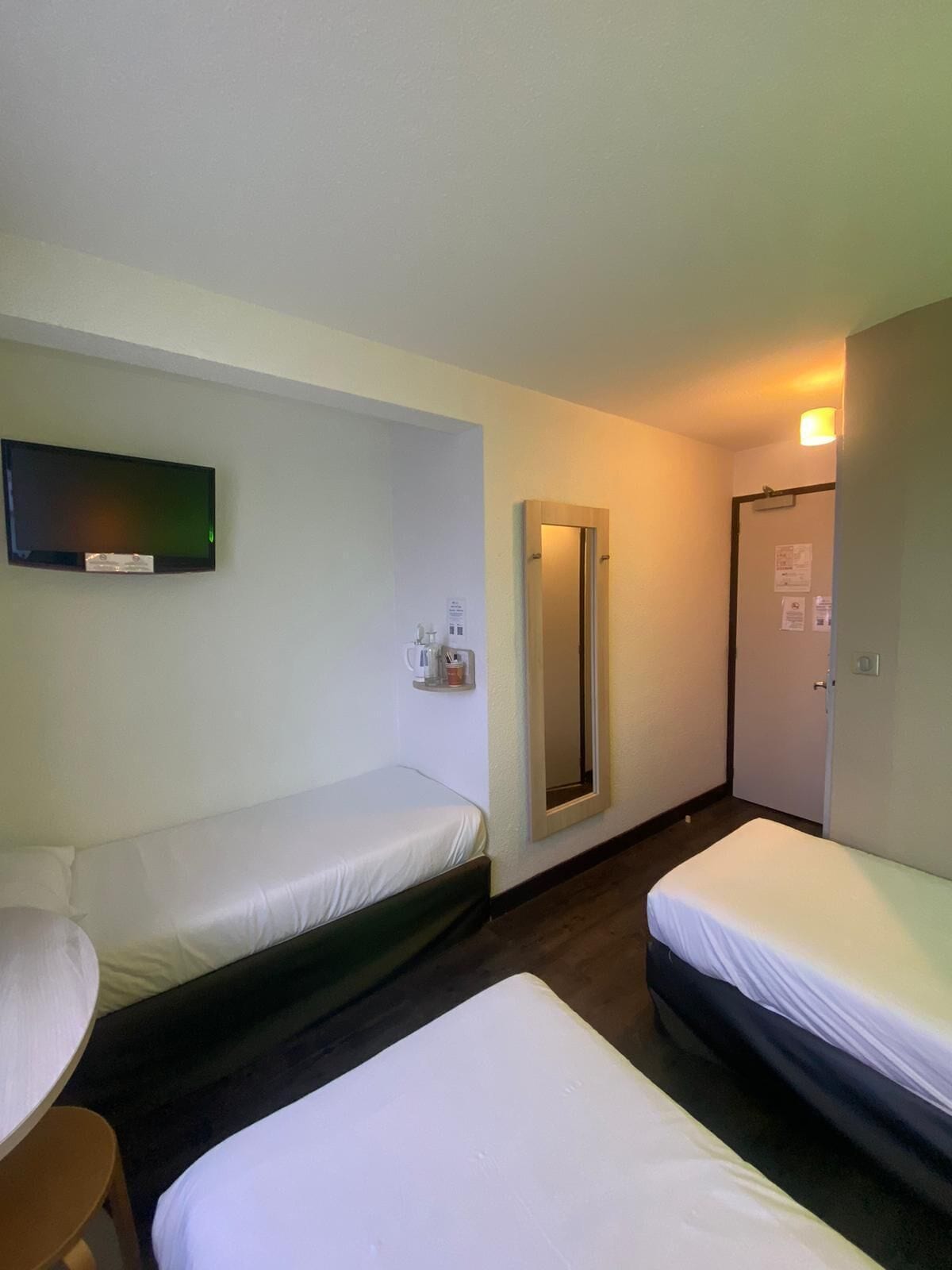 A hotel room with two single beds, a flat-screen TV mounted on the wall, a mirror, and a door with a light switch.