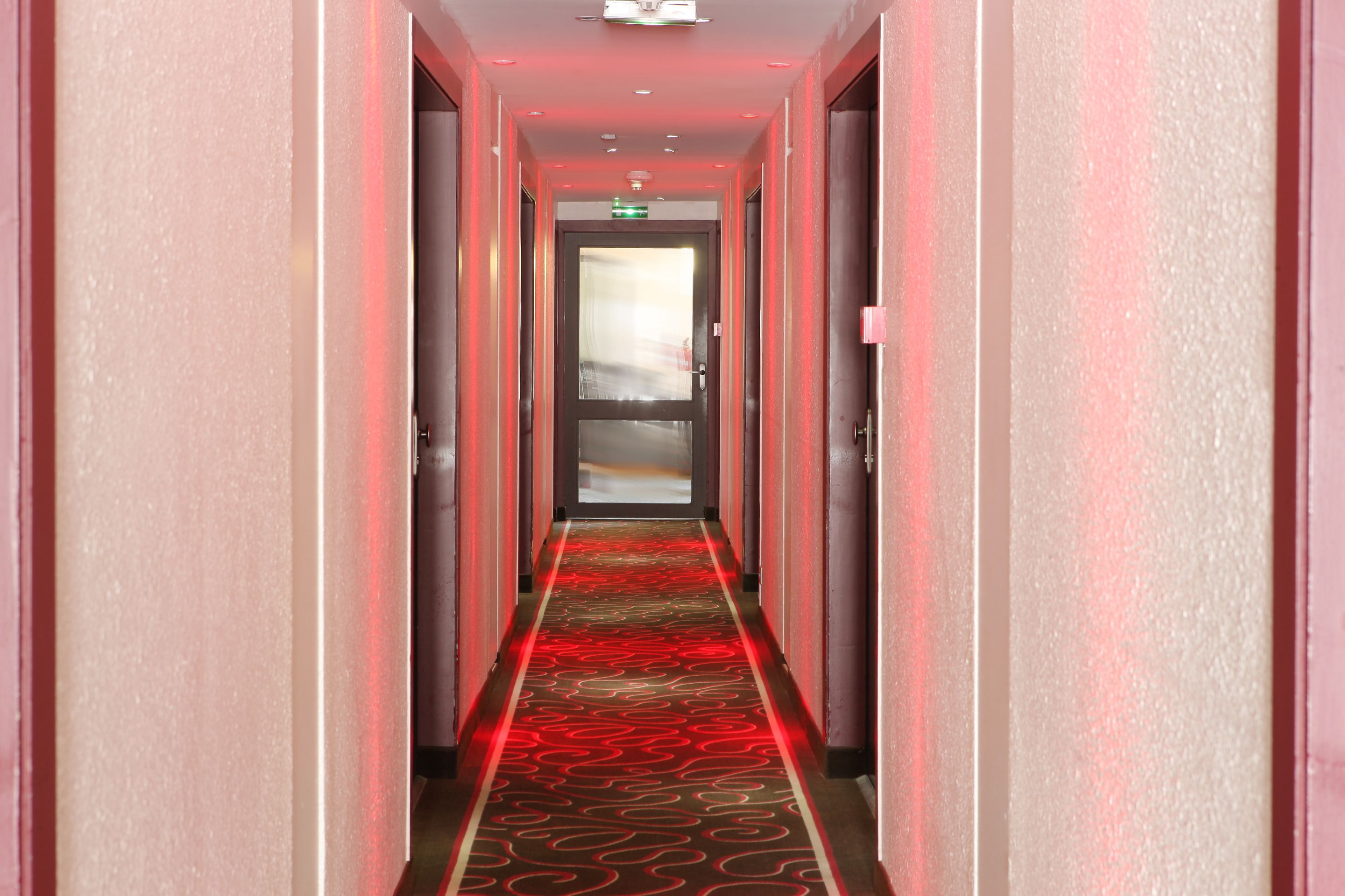 A hotel hallway with a red carpet and a glass door at the end.