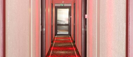 A hotel hallway with a red carpet and a glass door at the end.