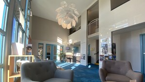 Eine großzügige Lobby mit hoher Decke, einem modernen Kronleuchter und einem blauen Teppich. Zwei graue Sessel und ein Holztisch mit Broschüren sind vorhanden.