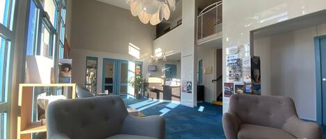 A spacious lobby with a high ceiling, a modern chandelier, and a blue carpet. There are two grey armchairs and a wooden table with brochures.
