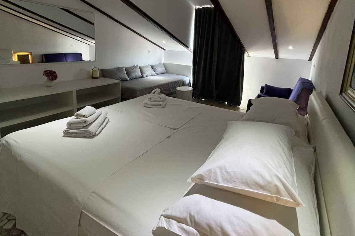 A modern hotel room with a large bed, a seating area with a sofa and armchairs, a small table, and a mirror above the bed.