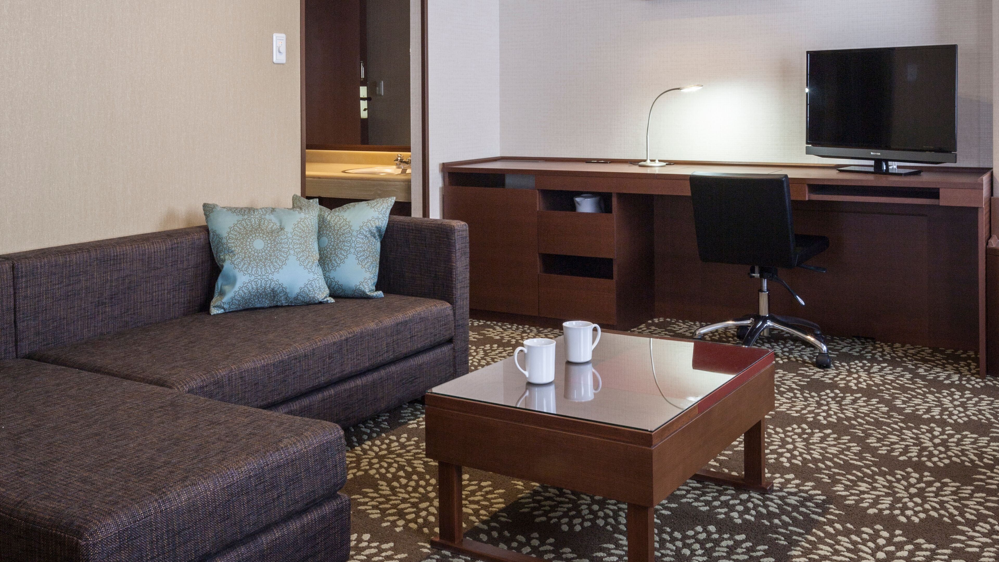 Habitación de hotel con sofá, una pequeña mesa con dos tazas, escritorio con computadora y televisor, y alfombra estampada.