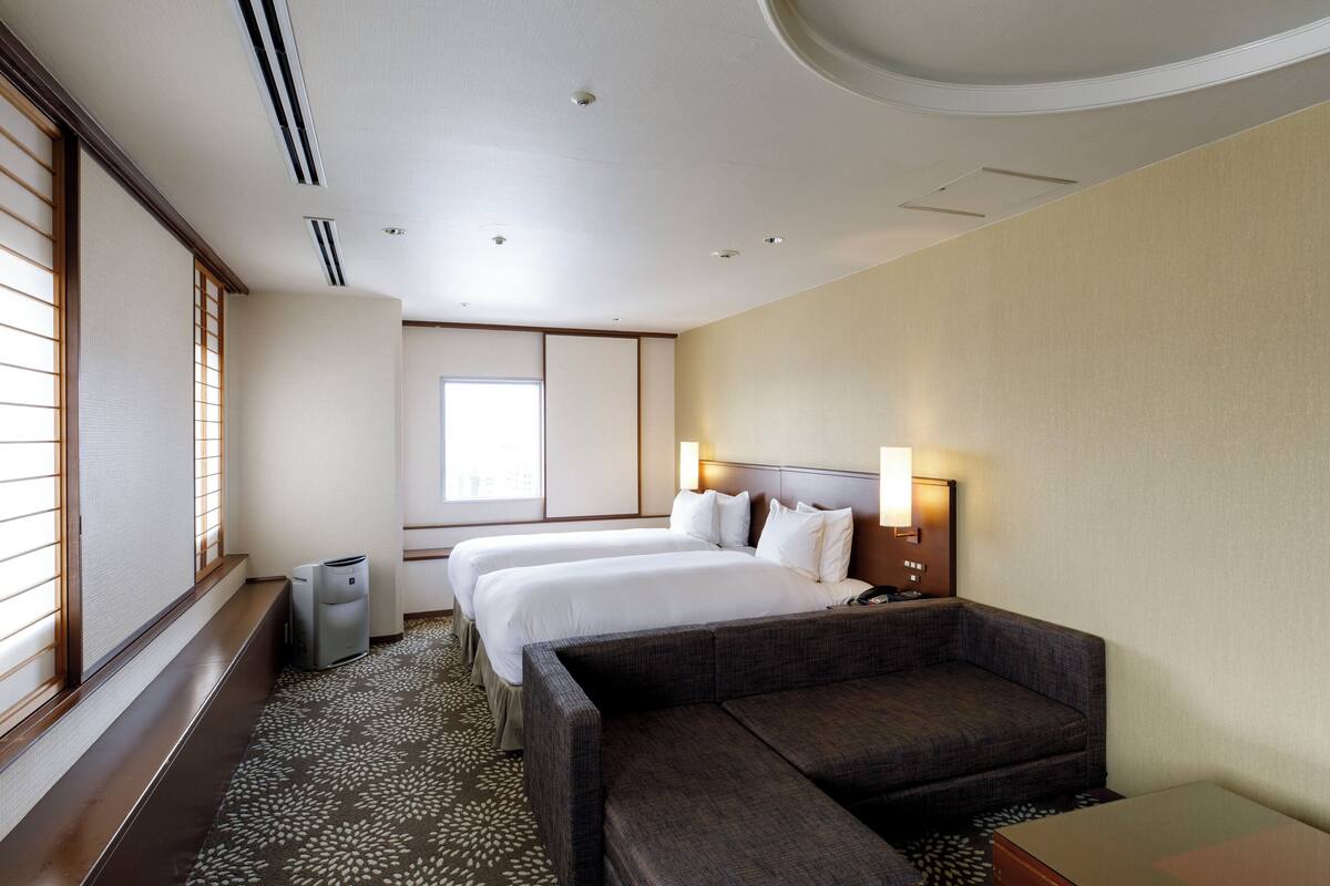 A hotel room with two beds, a sofa, a trash bin, and a window with blinds.