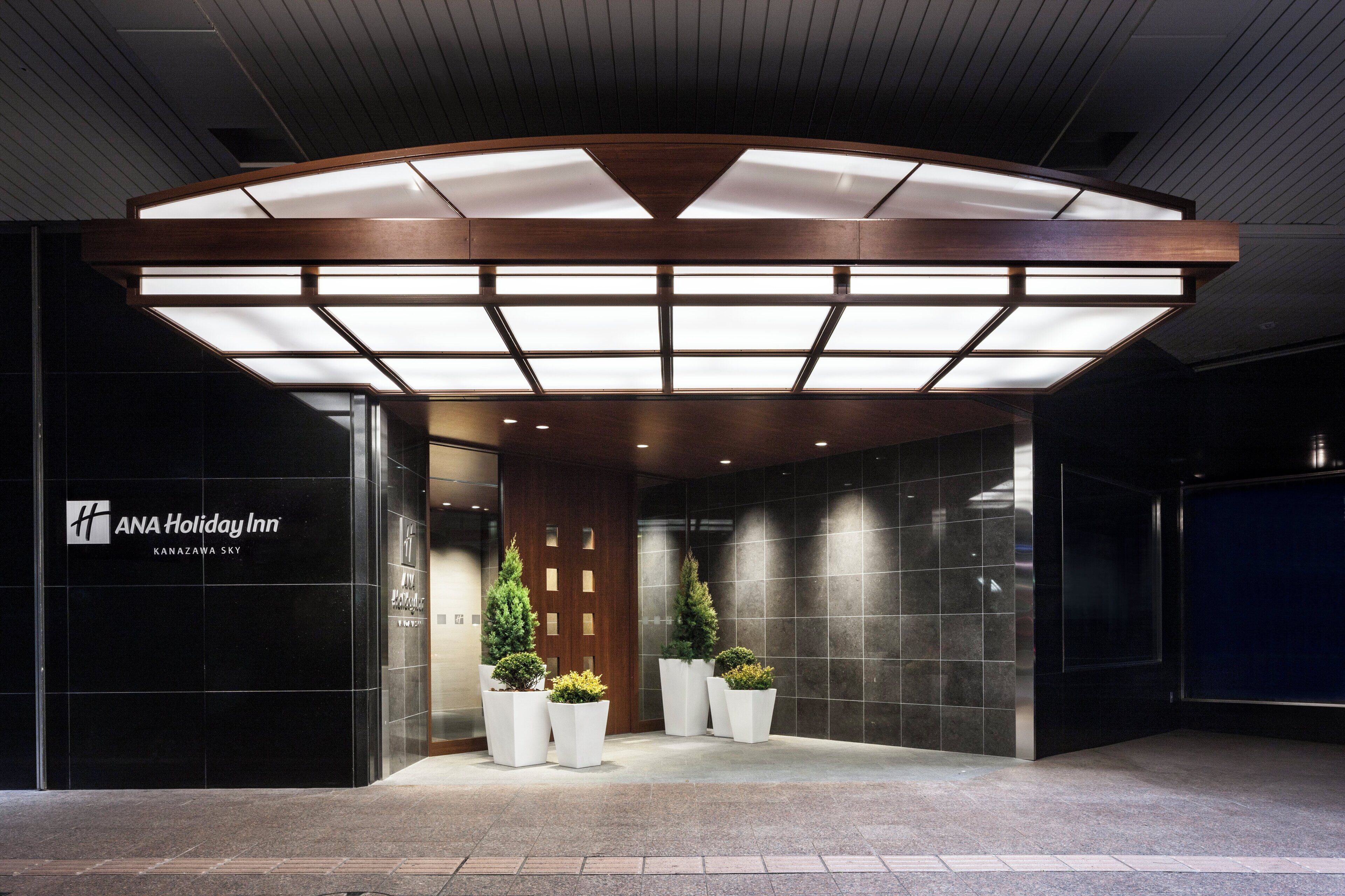 The entrance of ANA Holiday Inn with a modern design, featuring a large illuminated canopy, a reception desk, and potted plants.