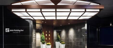 La entrada del ANA Holiday Inn con un diseño moderno, que presenta un dosel iluminado grande, un mostrador de recepción y plantas en macetas.