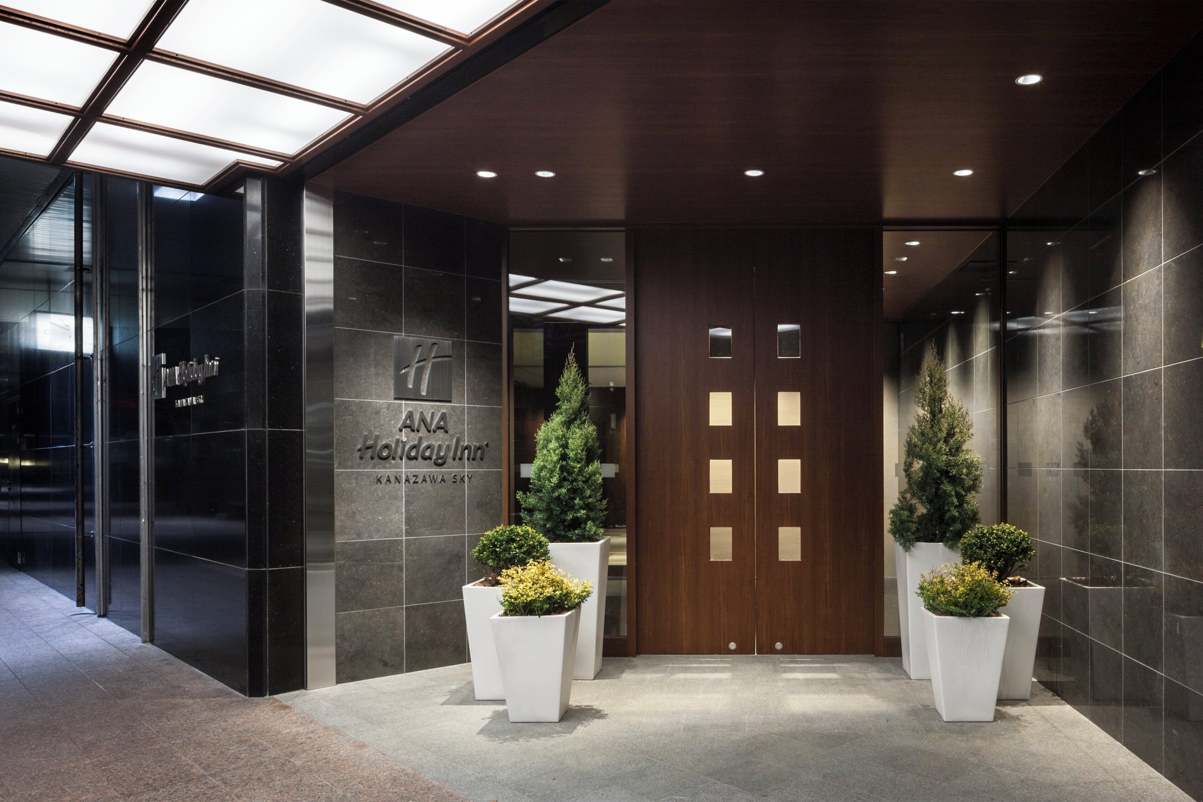 A hotel entrance with a modern design, featuring a glass facade, a wooden door, and potted plants.