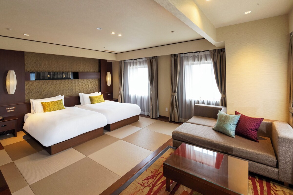 Habitación de hotel con dos camas, un sofá, un escritorio y un ventanal con cortinas.