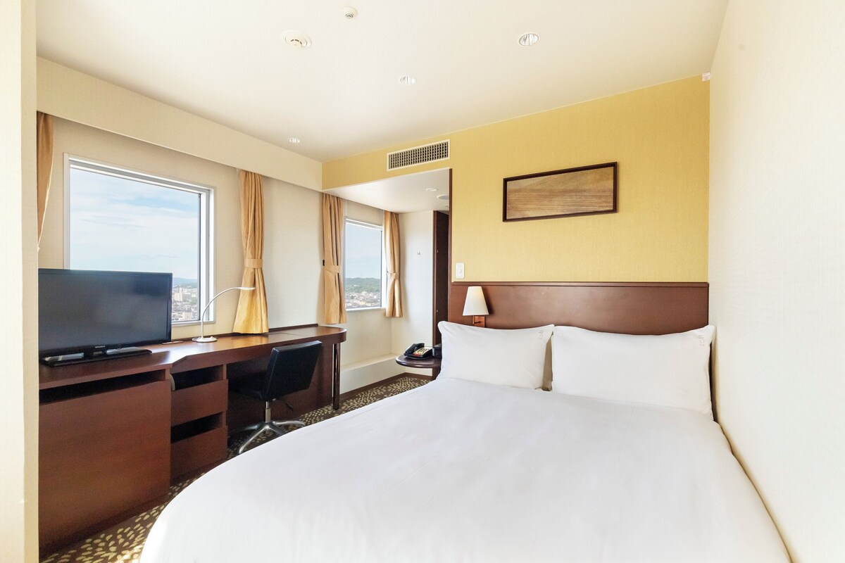 Habitación de hotel con una cama grande, un escritorio con televisor, teléfono y ventana con cortinas.