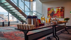 Una mesa de desayuno con cafeteras, tazas y una bandeja de fruta en una habitación moderna con una escalera y un ventanal grande.