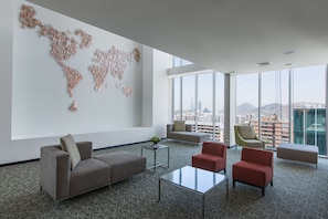 A modern hotel room with a large world map on the wall, a city view, and a mix of seating options including a sofa, chairs, and an ottoman.