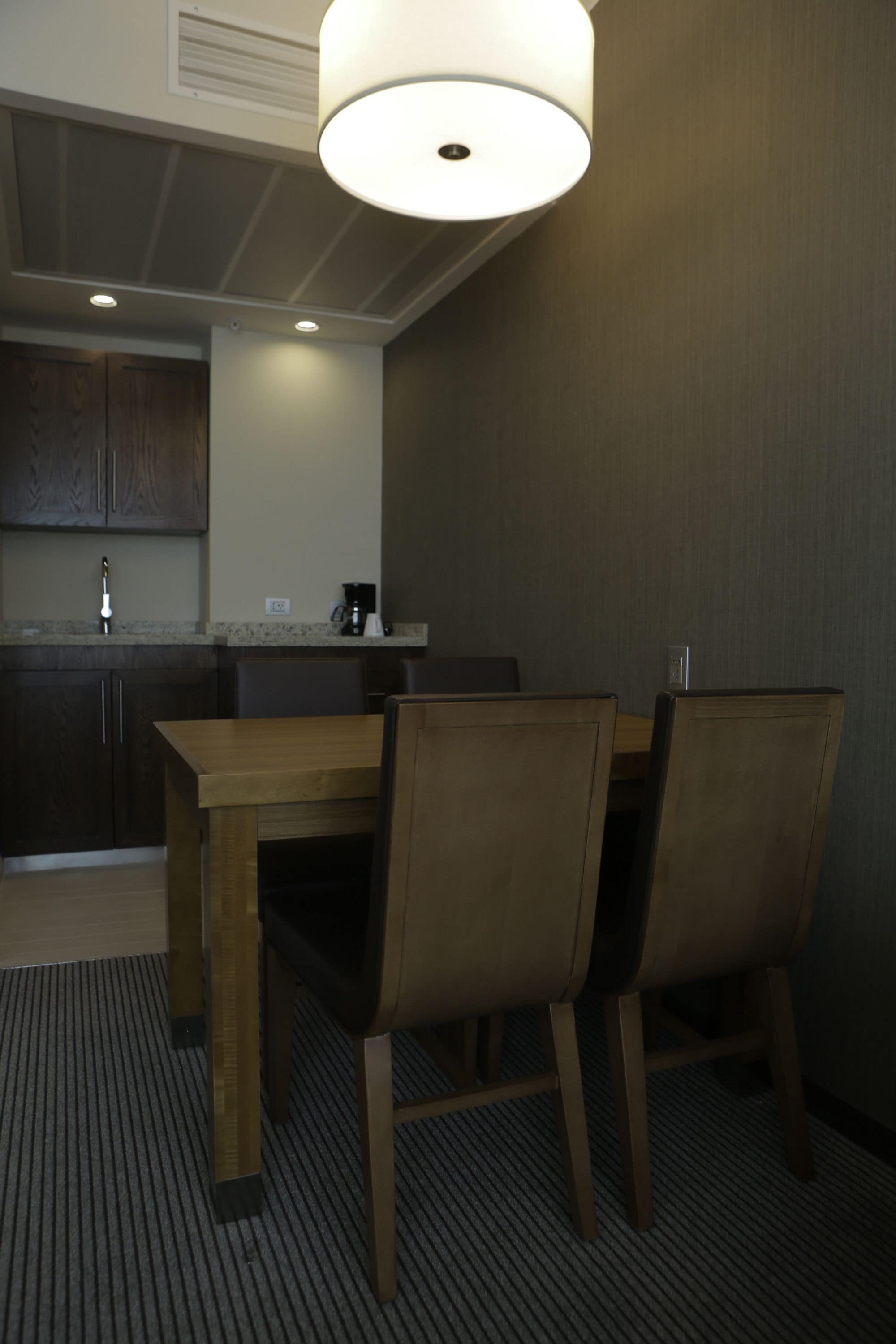Uma área de refeições com mesa e cadeiras de madeira, cozinha ao fundo e luminária no teto.
