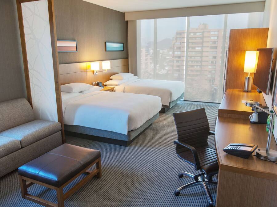 Habitación de hotel con dos camas, un sofá, un escritorio con lámpara y un amplio ventanal con vista a la ciudad.