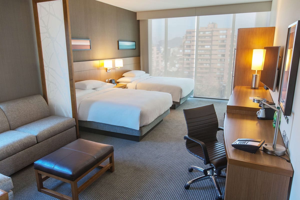 Ein Hotelzimmer mit Sofa, zwei Betten, einem Schreibtisch mit Lampe und Blick auf Gebäude.