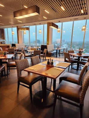 Ein modernes Restaurant mit hohen Decken, großen Fenstern und Blick auf die Stadt. Tische mit Stühlen sind angerichtet, und der Raum wird durch Pendelleuchten gut beleuchtet.