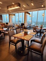 L’intérieur de ce restaurant moderne se caractérise par de hauts plafonds, de grandes fenêtres et une vue sur la ville. Les tables sont disposées avec des chaises, et l’espace est bien éclairé grâce à des luminaires suspendus.