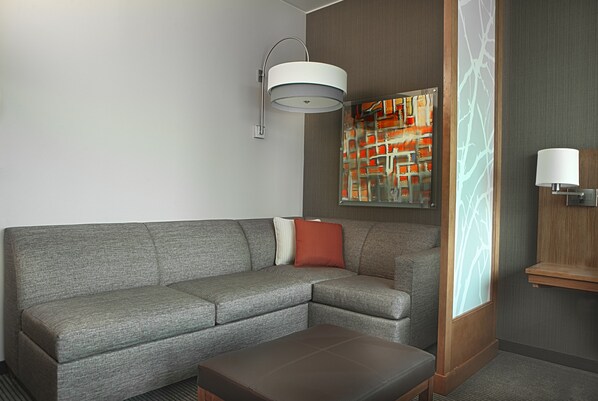 Ein modernes Hotelzimmer mit grauem Ecksofa, einem Holznachttisch, einer Stehlampe und einer dekorativen Wandverkleidung.